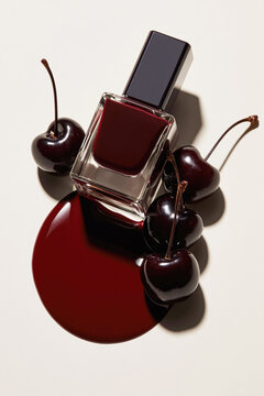 Overhead still life of burgundy nail polish surrounded by dark cherries and a glossy spill on a pale background.