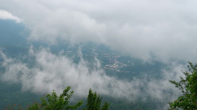 Stunning aerial vista of white fluffy clouds floating over a lush green valley, partially revealing a small town nestled among the mountains, Tkibuli Georgia