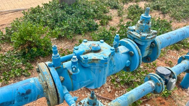 Blue water pipe and industrial valve installed beside a sandy landscaped area in an urban outdoor setting
