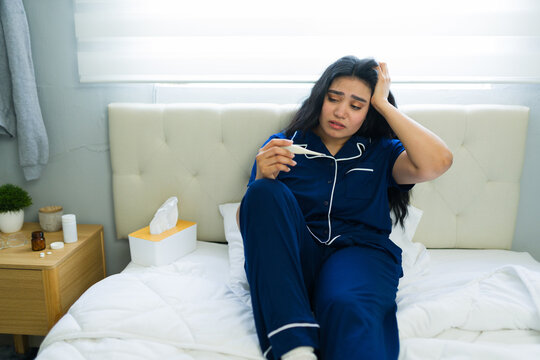 Sick woman feeling unwell in pajamas, holding her head and reading a digital thermometer showing high fever in bed