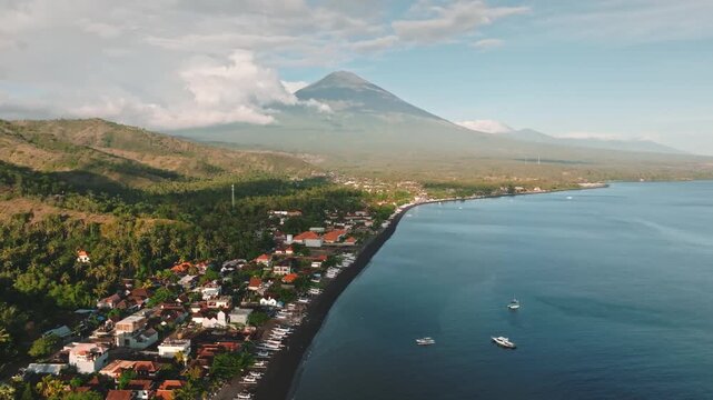 Amed village coastal landscape, traditional fishing boats on black sand beach with houses nestled among tropical trees and mount Agung volcano rising in background under cloudy sky. Aerial view shot