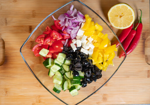 Vegetable salad in a bowl with sliced yellow bell peppers, red tomatoes, onions, cucumbers, olives, and feta cheese. Ingredients for a healthy salad, seasonal vegetables, chili peppers and a lemon.