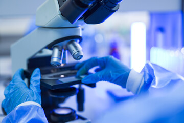 Gloved hands of a laboratory researcher turning the stage knob beneath the turret in a contemporary biomedical research facility, with blue reagent close at hand. © ultramansk