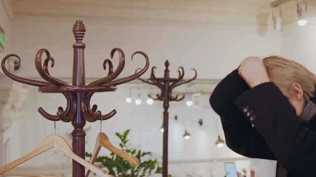 Close up of man trying on black hat in store examining fit and appearance. Wooden coat rack with hooks holds new headwear while man adjusts it to check comfort or style against his outfit