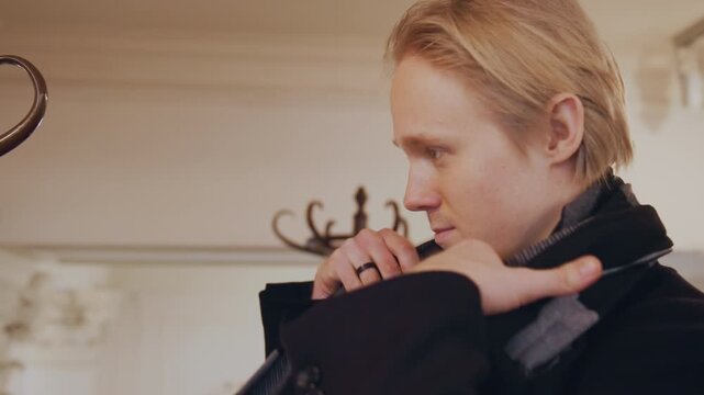 Close up of man adjusting his scarf in indoor setting with soft lighting. subject is wearing dark coat and background features ornate wall decor and decorative hooks. scene focuses