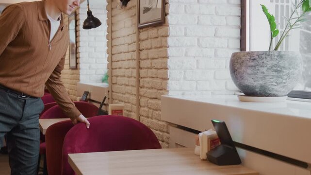 Man in brown shirt sits at table then stands up and moves to push back chair. setting is indoor cafe with white brick walls red chairs hanging lamps framed pictures on wall natural light