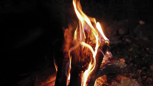 Close-up view of a bonfire burning brightly at night in a natural setting, with bright orange and yellow flames flickering intensely over the logs, creating a warm and cozy atmosphere