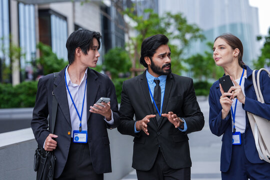 Three young business professionals wearing conference badges continue a discussion while walking past the building on the open terrace of a modern corporate campus in the city centre
