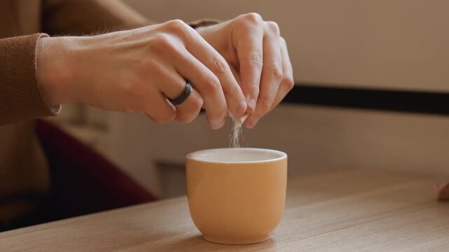 Close up of person hands squeezing sugar from small packets into orange ceramic cup on wooden table. action is performed steadily with fine details like ring worn and texture of skin