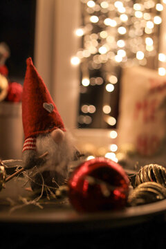 Cute Scandinavian Christmas Gnome Plush Toy in Red Pointed Hat with Golden Bokeh Lights, Baubles, and Garland Background