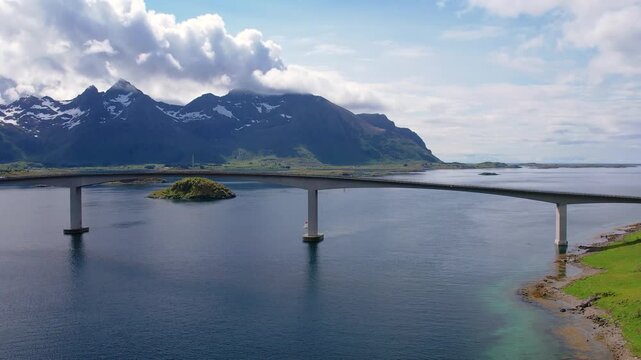 Drone aerial footage of a large bridge spanning the water in the Lofoten Islands, framed by mountains. The setting shows clear skies and nature surrounding the area