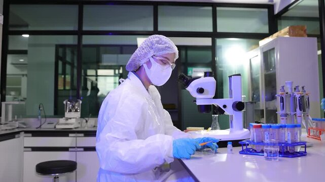 Scientist in protective suit using a microscope in a laboratory. Female scientist wearing a protective suit and mask working with a microscope in a laboratory. Medical research and development concept