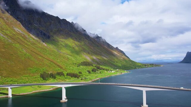 Drone aerial footage of a large bridge spanning the water in the Lofoten Islands, framed by mountains. The setting shows clear skies and nature surrounding the area