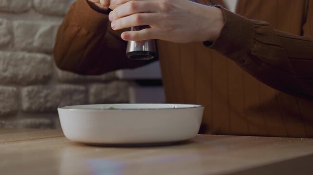 Close up of hands using metal pepper grinder to season food in white bowl on wooden table. Background features stone wall and soft indoor lighting emphasizing action of grinding pepper