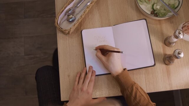 Woman writing mathematical equations in notebook at wooden table with soft lighting. scene includes open book displaying handwritten formulas surrounded by objects like salad bowl and salt