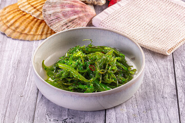 Fresh green seaweed salad in ceramic bowl on weathered wood, with scallop shells and textured cloth background © Andrei Starostin