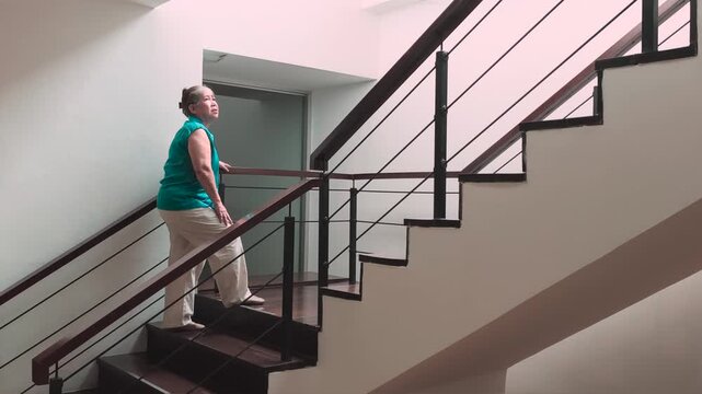 60s old senior asian woman slowly climbing staircase with knee joint pain or leg muscle atrophy, indoor home scene during daytime, health and body care, A11Y, and elderly mobility concept