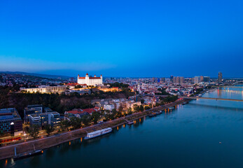 Obraz premium Bratislava skyline at dusk with Danube River and city lights visible. City lights shine along the Danube River in Bratislava during dusk. The castle is on the hill.
