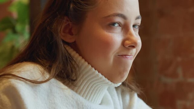 Close up of young woman with long brown hair wearing white turtleneck sweater showing subtle facial expressions and slight head movements. background features warm indoor lighting