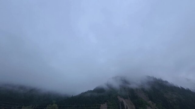Timelapse of heavy low hanging clouds and thick fog rolling rapidly over a dark evergreen forested mountain ridge under moody overcast skies