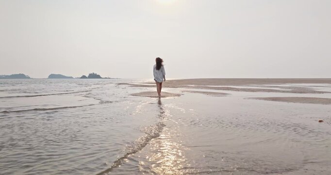 Woman in casual attire walking barefoot across an exposed sandbar at low tide, with the sun reflecting on shallow water and distant islands completing a serene tropical landscape. Ko Lanta, Thailand