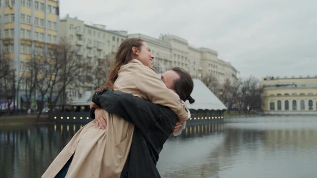 Close up of man and woman embracing by lake with city buildings in background. couple interacts affectionately including hugging and playful gestures like touching hair against overcast sky