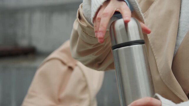 Close up of person holding and opening stainless steel thermos revealing inner lid. person wears light colored clothing with visible texture details in outdoor setting with soft lighting