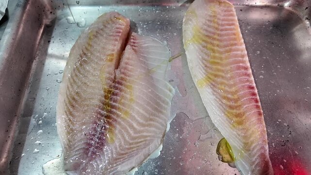 Two raw tilapia fish fillets are displayed in a metal container. The fillets are white and pink with visible muscle fibers and are slightly moist