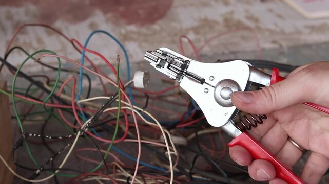 An electrical tool for stripping wires (a stripper) in a craftsman's hand, close-up. A hand with metal pliers-strippers over a pile of tangled cables. Hand tools