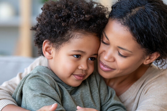Mother hugs her young child tenderly. Her eyes show gentle affection. The child rests close, looking calm and content. Soft indoor lighting enhances the warmth of the moment