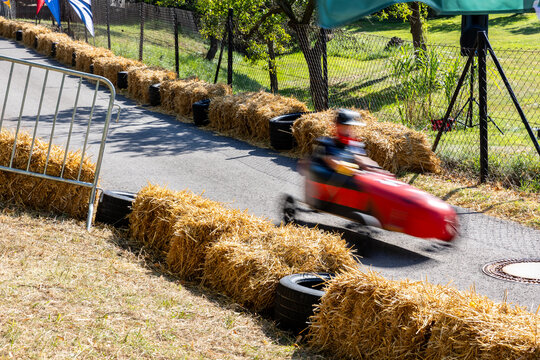 Soap box racer speeds through curved street course in Germany, captured with motion blur beside hay bales and tire barriers. Summer race conveys speed, competition, excitement and festival atmosphere