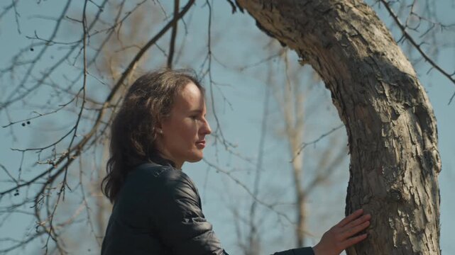 Close up of woman touching rough bark of tree outdoors on clear day. Her long hair moves slightly in wind wearing dark jacket and blue shirt background shows bare branches against sky
