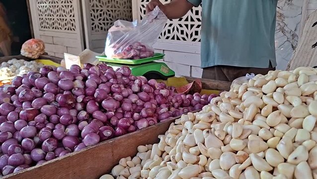 fresh red shallots being packed into plastic bag by market seller on scale with large piles of organic garlic cloves and red onions displayed on wooden table at traditional Asian produce market 