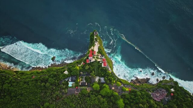 Uluwatu temple perched on a steep Balinese cliff, overlooking the Indian ocean with lush greenery and powerful waves crashing against rugged coastline, dramatic scenic seascape. Aerial top down view