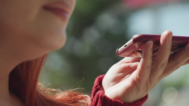 Close up of woman with red hair holding and using smartphone outdoors. Woman wears textured knit sweater her hand positioned to interact with device against soft natural light and blurred
