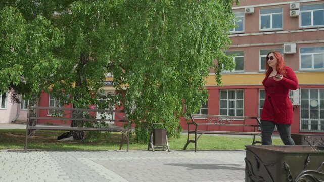 Woman in red sweater walks through outdoor urban park area holding her phone and purse. scene features green trees metal benches decorative planters with leaf patterns paved ground grassy