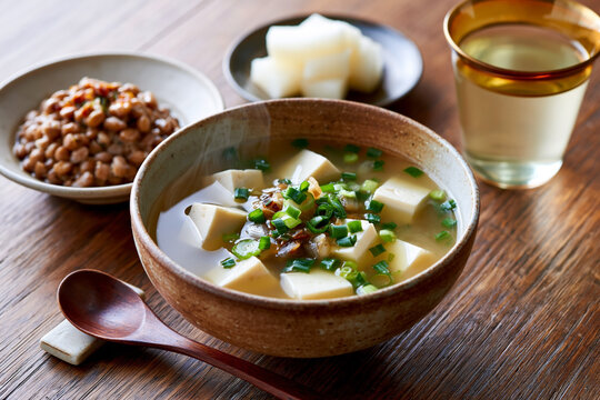 Bowl of miso soup with tofu and green onions served alongside natto and pickled vegetables on a wooden table with a glass of tea