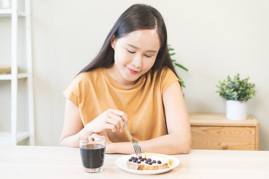 Happy young Asian woman smiling while enjoying a healthy breakfast with banana and blueberry toast with cream cheese, served with hot black coffee on a wooden table in the morning at home.