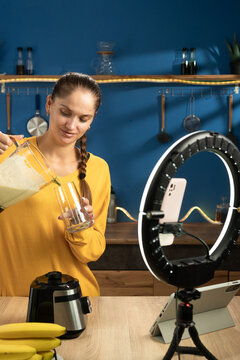 Female food blogger prepares a fruit smoothie in her home kitchen while recording content with a smartphone and ring light for social media followers.