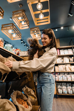Happy female pet owner holding her fluffy brown dog while choosing healthy treats and supplies from wooden containers in a modern specialty boutique.
