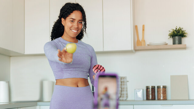 Smiling African American Woman In Her 20s, Holding A Green Apple While Taking A Photo At Home In The Kitchen. Embracing Healthy Living And Nutrition. Weight Loss Concept.