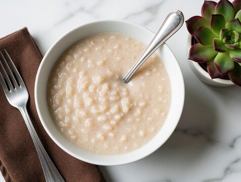 Hot cream of wheat porridge in white bowl with metal spoon. Healthy breakfast meal on marble table near brown napkin, succulent plant. Warm cooked cereal served for nutritious morning diet. Simple