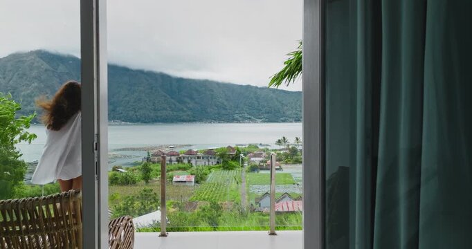 Woman on a hotel balcony admires the tranquil panorama of lake Batur and volcanic mountains in Bali, enjoying a peaceful tropical getaway and reflective moment during her vacation