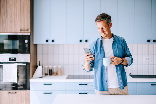 Smiling Man in Modern Kitchen Checking Phone and Drinking Coffee &mdash; Casual Morning Home Routine for Remote Work