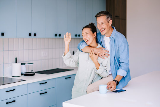 Happy Couple Hugging and Waving in Modern Kitchen While Drinking Coffee and Laughing Together
