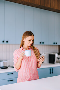 Woman In Modern Kitchen Checking Smartphone While Holding Coffee Mug &mdash; Casual Remote Work Morning Routine