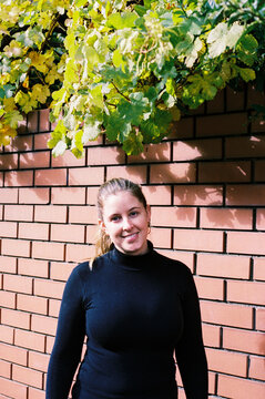 Woman Smiling Under Green Foliage on Brick Wall