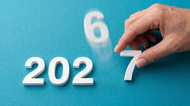 Hand placing a white number 7 next to 202 to change the year from 2026 to 2027 on blue.