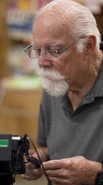 An elderly man is shopping in a grocery store. He examines items on the shelf with care. The store is busy with other customers.
