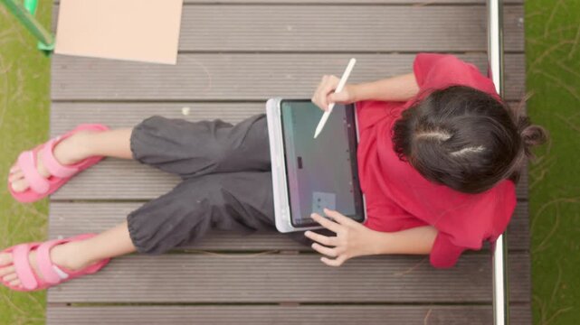 Top view of an Asian kid enjoying a relaxing hobby, drawing digital art on a tablet. Selective focus captures the elementary student spending casual leisure time at an outdoor school.